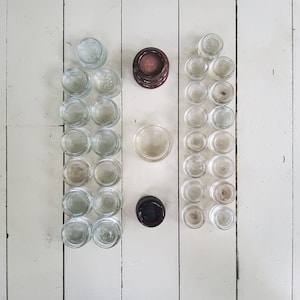 May include: Overhead view of small, clear glass jars with lids, arranged in rows on a white wooden surface. Also present are two dark brown and one black jar with lids, and one clear lid. The jars are empty.
