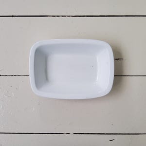 May include: A small, rectangular white dish with rounded corners. The dish is empty and sits on a white, weathered wooden surface with visible wood grain. The dish is likely for holding small items.