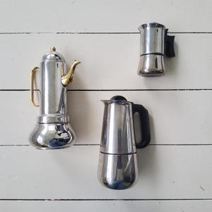 Op de afbeelding: Drie roestvrijstalen espressomakers worden getoond tegen een witte achtergrond met horizontale planken. De espressomakers variëren in grootte en ontwerp, met zwarte handvatten en tuiten. Eén heeft een goudkleurig handvat en tuit.