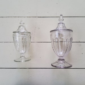Può includere: Due barattoli di caramelle in vetro trasparente con coperchi. I barattoli hanno la forma di urne con una base larga e un collo stretto. I barattoli sono vuoti e hanno un aspetto vintage.
