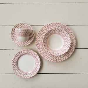 May include: A white teacup, saucer, and plates with a delicate red floral pattern. The set includes a teacup, a saucer, a small plate, a medium plate, and a large plate. The plates and cup are arranged on a white wooden surface.