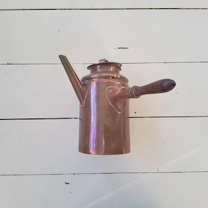 May include: A copper tea kettle with a wooden handle and spout. The kettle has a heart-shaped detail on the side and a lid on top. The copper has a shiny, reflective surface.