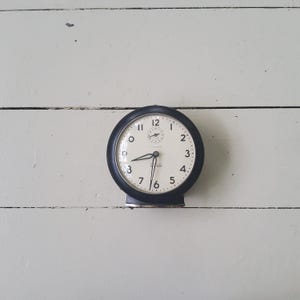 May include: A vintage black alarm clock with a white face. The clock has a round shape with a black bezel and a small base. The clock face features black numerals and hands, with the time displayed as 8:20.