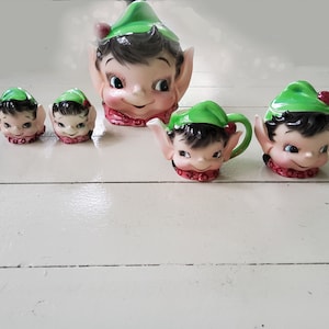 May include: A collection of vintage ceramic elf-themed teaware. The set includes a teapot, creamer, sugar bowl, and two small cups, all featuring elf faces with green hats, black hair, and rosy cheeks. The teapot is the largest piece.