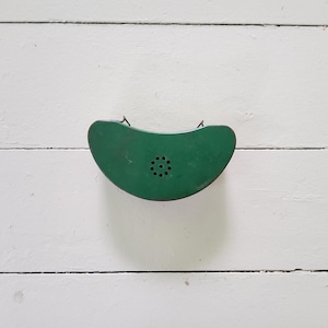 May include: A vintage, green metal wall-mounted telephone receiver. The receiver has a curved shape with a series of small holes in the center and two metal loops for hanging. The object is set against a white, wooden background.