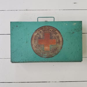 Op de afbeelding: Vintage turquoise metalen EHBO-kit met een handvat. De kit heeft een cirkelvormig embleem met een rood kruis en de woorden "Safety Every Industry". De doos heeft een rechthoekige vorm en een scharnierend deksel.