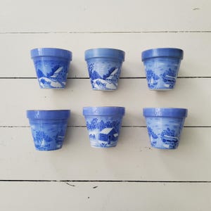 Può includere: Sei piccoli vasi di fiori in ceramica blu con un design paesaggistico. Ogni vaso presenta una dettagliata illustrazione bianca e blu di una casa, alberi e uno specchio d'acqua. I vasi sono disposti su una superficie di legno bianco.