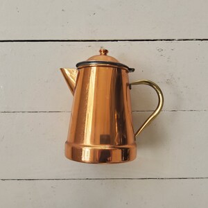 May include: A copper coffee pot with a brass handle and a lid. The pot has a shiny, reflective surface.