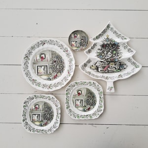 Pode incluir: Um conjunto de pratos e um prato de servir com tema natalino. Os pratos são brancos com uma borda de folhas de azevinho verdes e vermelhas. O centro de cada prato apresenta uma cena de árvore de Natal e lareira. O prato de servir tem a forma de uma árvore de Natal.