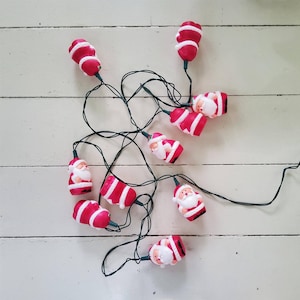 May include: A string of festive lights featuring multiple small, moulded plastic Santa Claus figures. Each figure is painted in red and white, attached to a thin, dark green electrical cord. The Santa figures are arranged in a tangled cluster.