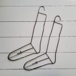 May include: Two dark metal sock drying forms shaped like feet with a hook at the top for hanging. Each frame is constructed from thin wire and features a horizontal support bar across the foot section with a small sliding adjustment mechanism.