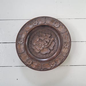 May include: A round, dark brown wooden plate with intricate floral carvings. The plate features a central floral design surrounded by a border of leaves and flowers. The wood grain is visible, adding to the rustic charm of the decorative plate.