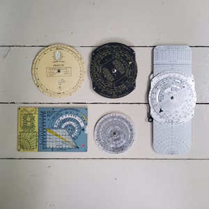 May include: Five circular and rectangular navigation tools with dials and markings. The tools are made of metal and plastic and have a vintage look. The tools are arranged on a white surface.