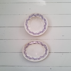 May include: Two white ceramic plates with a purple floral border. One is a shallow bowl, and the other is a flat plate. The plates are arranged on a white, weathered wooden surface.
