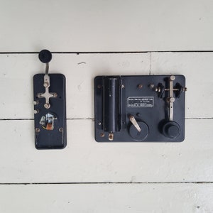 Könnte beinhalten: Zwei schwarze Vintage-Radiokomponenten. Die größere Komponente hat einen silbernen Knopf und den Text "REXREX PRACTICE AIR-GAP TYPE" aufgedruckt. Die kleinere Komponente hat einen silbernen Knopf und einen silbernen Draht.