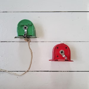 May include: Two vintage metal cord winders. One is green, the other is red. Both have a crank handle and a small metal base. The winders are mounted on a white, wooden surface.