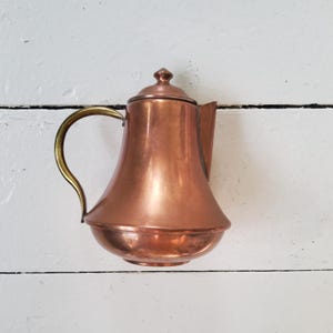 May include: A copper teapot with a brass handle and a lid. The teapot has a rounded base and a curved spout. The handle is a contrasting gold color. The teapot is likely used for brewing tea or coffee.