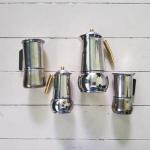 May include: Four stainless steel stovetop espresso makers arranged together. Two pots feature gold-toned handles and lid knobs, while the other two have black handles. Each coffee maker has a cylindrical body, a hinged lid, and a side handle.