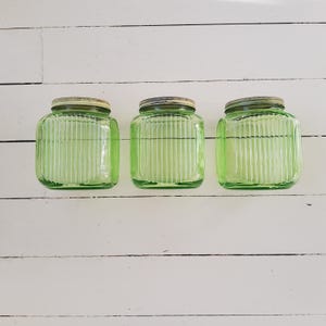Könnte beinhalten: Drei grüne Glasbehälter mit geriffelten Seiten und silbernen Deckeln. Die Behälter sind in einer Reihe auf einer weißen Holzoberfläche angeordnet.