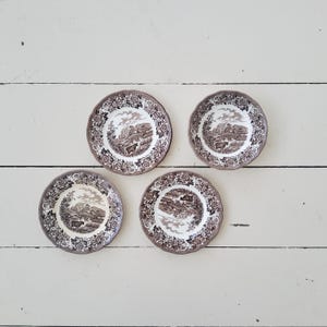 Può includere: Quattro piatti decorativi vintage marroni e bianchi con un design paesaggistico. Ogni piatto presenta un bordo smerlato e una scena dettagliata di un edificio, alberi e animali. I piatti sono disposti su una superficie di legno bianco.