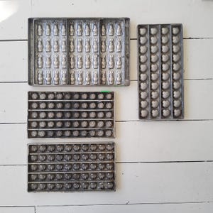 May include: A collection of vintage metal molds for making candies or chocolates. The molds are in various shapes, including figures and seashells. The molds are arranged on a white wooden surface.