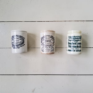 Pode incluir: Três frascos de cerâmica branca com etiquetas azuis e pretas. Os frascos contêm marmelada de laranja. Os rótulos dizem "James Keiller & Sons, Dundee, Marmelada de laranja, Medalha para marmelada 1868", "James Keiller & Son Ltd, Dundee, Marmelada de laranja, 12 Medalha 1868, Dundee, Escócia", e "James Keiller & Son Ltd, Estabelecido em 1871, Dundee original, Marmelada de limão em pedaços grossos, 12 fl oz 341 ml, Produto da Escócia".