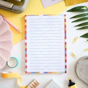 May include: A white notepad with lined paper and a colourful border. The notepad is on a white surface with a pink pencil, a yellow ruler, a yellow and blue tape roll, a yellow ribbon, a grey stone coaster, a grey and white stone coaster, and a yellow highlighter.