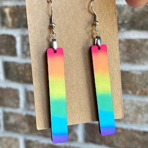 Könnte beinhalten: Ein Paar regenbogenfarbene Gradienten-Ohrringe mit silbernen Haken. Die Ohrringe sind rechteckig und haben eine glatte, glänzende Oberfläche.