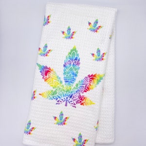 Może przedstawiać: Biały ręcznik kuchenny z dużym wzorem liścia konopi w stylu tie-dye w tęczy. Mniejsze liście konopi w stylu tie-dye są rozrzucone po ręczniku.