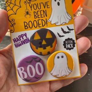 May include: A set of four Halloween themed button pins. The pins feature a ghost, a pumpkin, bats, and the word "BOO". The text "You've Been Booed!" and "Happy Halloween" are also included on the card. The card also says "Button Pack".