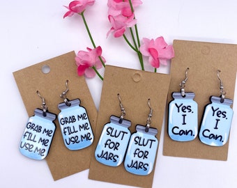 May include: Four pairs of earrings featuring mason jar-shaped charms with various sayings. The sayings include "Grab Me Fill Me Use Me", "Slut For Jars", and "Yes I Can".