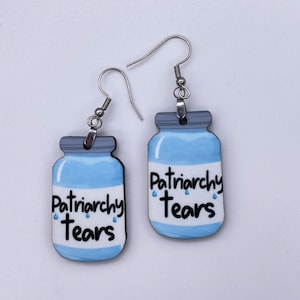 Op de afbeelding: Een paar oorbellen met een potontwerp. De oorbellen hebben een lichtblauwe body en een grijze deksel. De woorden "Patriarchy Tears" zijn gedrukt op een wit label, met blauwe traanaccenten. Zilverkleurige oorhaken.