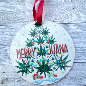 May include: A white oval Christmas ornament with a green marijuana leaf Christmas tree design. The tree is decorated with colorful lights and presents. The text "Merry Juana" is written on the ornament.