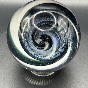 May include: A clear glass marble with a swirling design of white, black, and blue. The marble has a spiral pattern and is set against a dark background. The glass has a reflective quality.