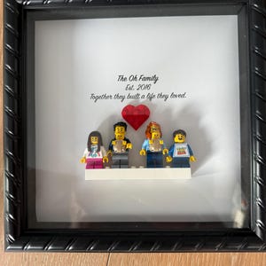 May include: A framed family portrait featuring Lego figures. The black frame surrounds a white background with the text "The Oh Family Est. 2016" and "Together they built a life they loved." Lego figures represent the family, with a red heart.