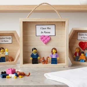 May include: Three light-colored wooden display frames containing plastic building block figures. The center square frame features a pink heart and the text I Love You to Pieces. The two hexagonal frames feature the text Custom Family to Pieces.