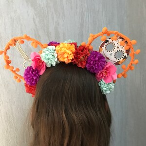 K&ouml;nnte beinhalten: Ein orangefarbenes Stirnband mit einem Totenkopf und Blumen. Das Stirnband ist mit orangefarbenen Bommeln und einem Zucker-Sch&auml;del verziert. Die Blumen sind rosa, lila, orange und gr&uuml;n.
