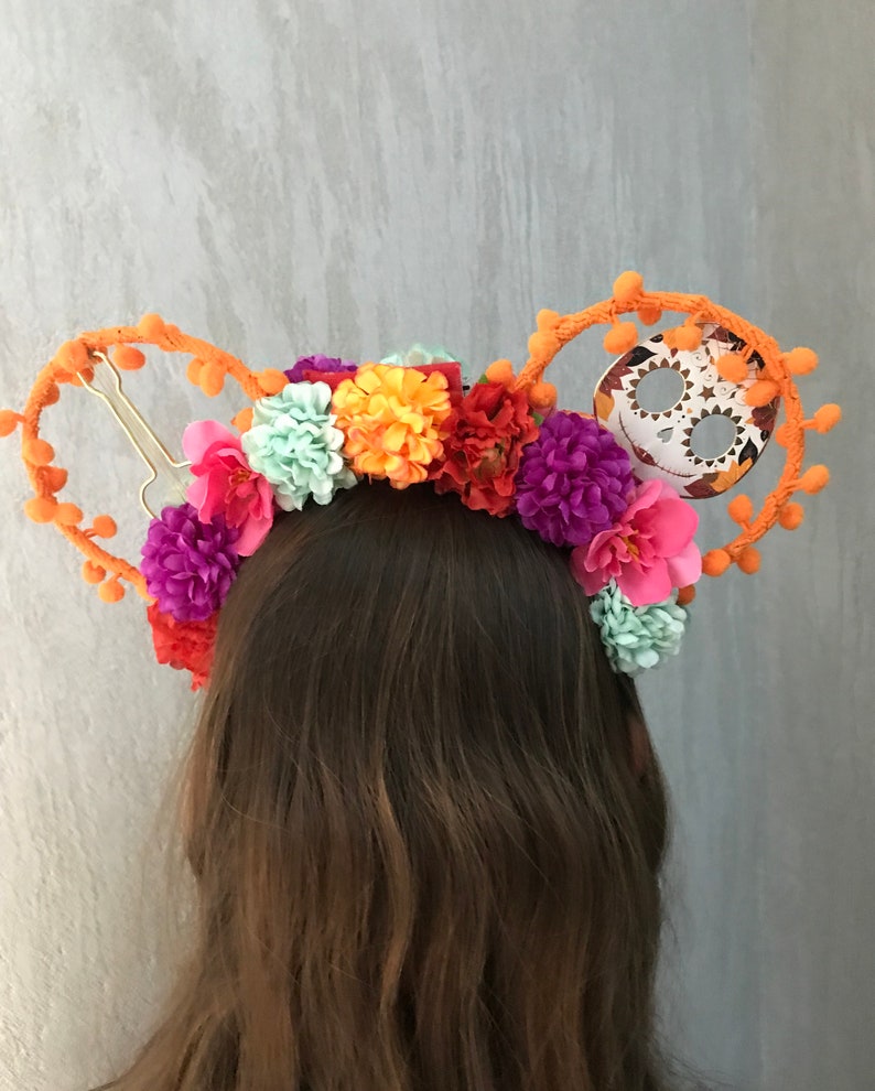 K&ouml;nnte beinhalten: Ein orangefarbenes Stirnband mit einem Totenkopf und Blumen. Das Stirnband ist mit orangefarbenen Bommeln und einem Zucker-Sch&auml;del verziert. Die Blumen sind rosa, lila, orange und gr&uuml;n.