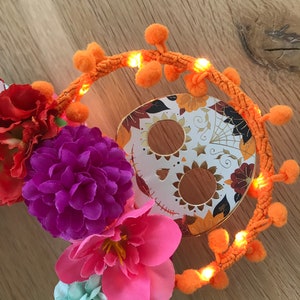 K&ouml;nnte beinhalten: Ein Dia-de-los-Muertos-Kranz mit einem Zucker Totenkopf, orangefarbenen Bommeln und bunten Blumen.