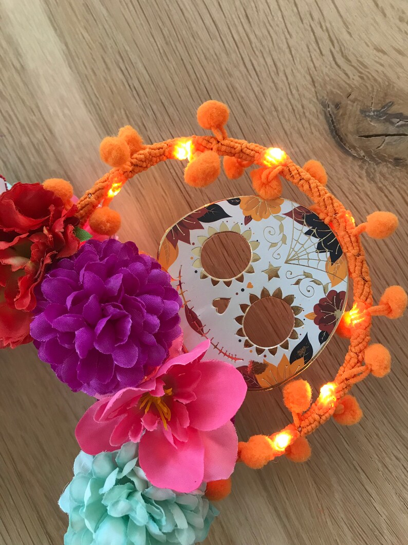 K&ouml;nnte beinhalten: Ein Dia-de-los-Muertos-Kranz mit einem Zucker Totenkopf, orangefarbenen Bommeln und bunten Blumen.