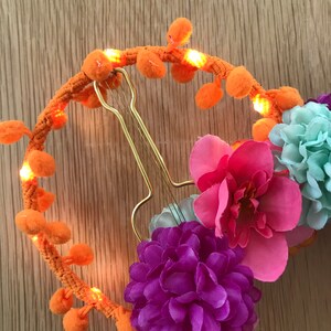 K&ouml;nnte beinhalten: Ein leuchtend orangefarbenes Stirnband mit Bommeln und kleinen Lichtern. Das Stirnband ist mit rosa, lila und blauen Stoffblumen verziert.