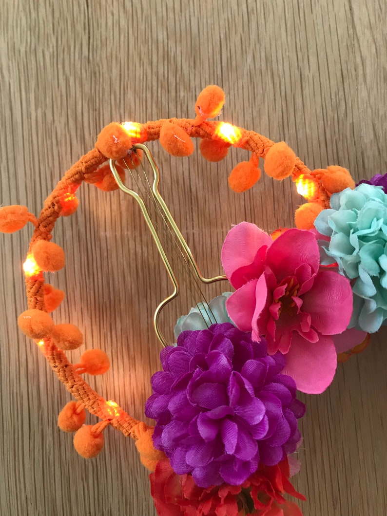 K&ouml;nnte beinhalten: Ein leuchtend orangefarbenes Stirnband mit Bommeln und kleinen Lichtern. Das Stirnband ist mit rosa, lila und blauen Stoffblumen verziert.