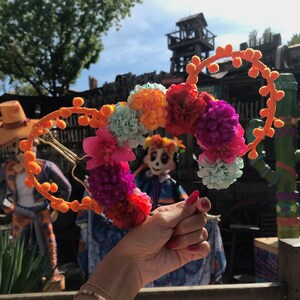 K&ouml;nnte beinhalten: Ein Paar orangefarbene und goldene Mickey Mouse Ohren mit einem Blumengirlanden aus rosa, lila, orange und blauen Blumen. Die Girlande ist kreisf&ouml;rmig und mit einem goldenen Draht an den Ohren befestigt.