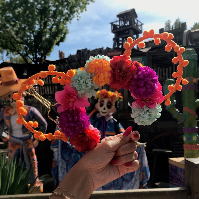 K&ouml;nnte beinhalten: Ein Paar orangefarbene und goldene Mickey Mouse Ohren mit einem Blumengirlanden aus rosa, lila, orange und blauen Blumen. Die Girlande ist kreisf&ouml;rmig und mit einem goldenen Draht an den Ohren befestigt.