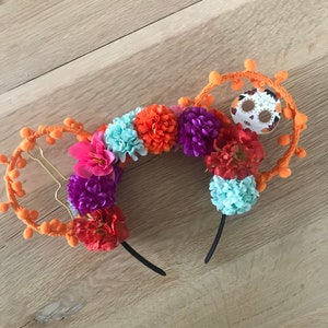 K&ouml;nnte beinhalten: Orangefarbene und schwarze Mickey-Maus-Ohren mit einem Totenkopf-Design und bunten Blumen. Die Blumen sind rosa, lila, orange und blau.