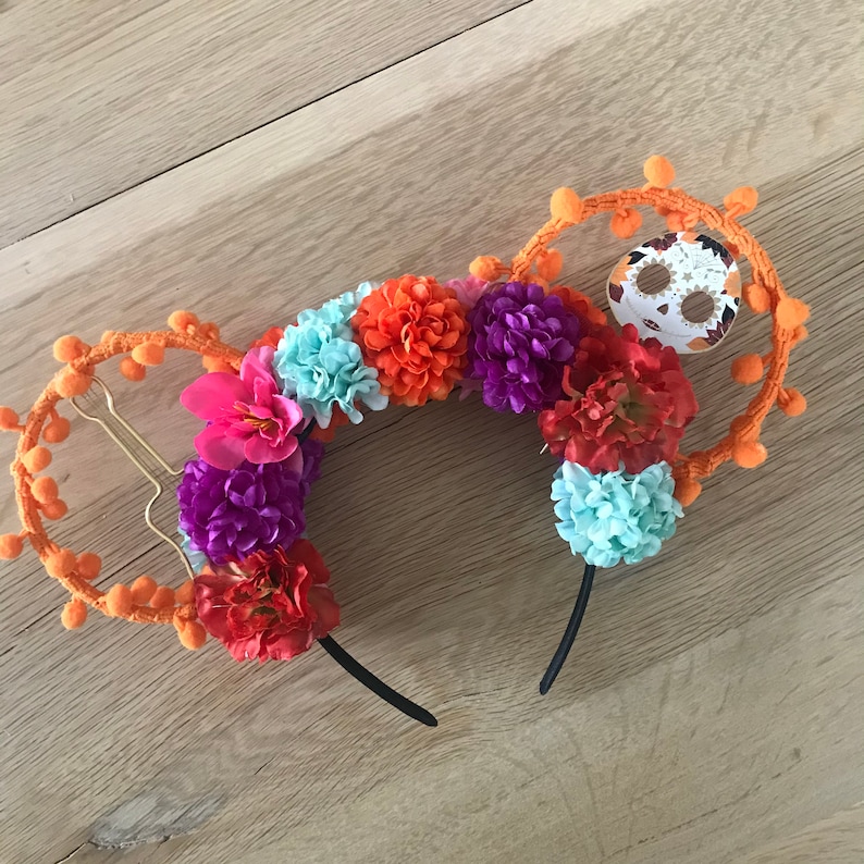 K&ouml;nnte beinhalten: Orangefarbene und schwarze Mickey-Maus-Ohren mit einem Totenkopf-Design und bunten Blumen. Die Blumen sind rosa, lila, orange und blau.