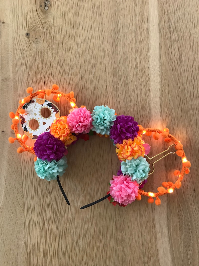 K&ouml;nnte beinhalten: Ein Paar leuchtende Minnie Mouse Ohren, verziert mit bunten Blumen und einem Totenkopf aus Zucker f&uuml;r den Tag der Toten. Die Ohren sind orange und haben eine Lichterkette aus orangefarbenen Lichtern am Rand. Die Blumen sind rosa, orange, lila und blau.