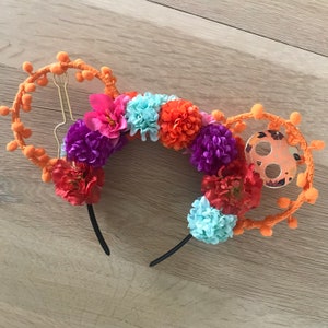 K&ouml;nnte beinhalten: Ein orange-schwarzes Stirnband mit Blumenmuster. Das Stirnband ist mit orangefarbenen Bommeln, lila, rosa, blauen und orangefarbenen Blumen verziert. Das Stirnband hat ein schwarzes Gummiband.