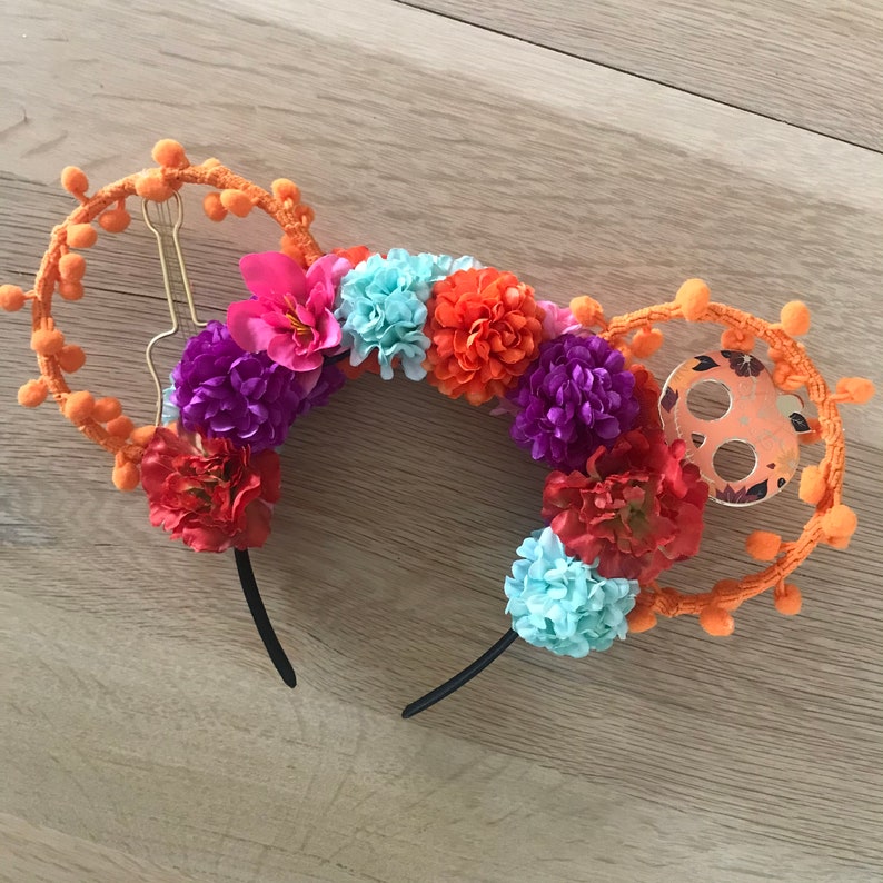 K&ouml;nnte beinhalten: Ein orange-schwarzes Stirnband mit Blumenmuster. Das Stirnband ist mit orangefarbenen Bommeln, lila, rosa, blauen und orangefarbenen Blumen verziert. Das Stirnband hat ein schwarzes Gummiband.