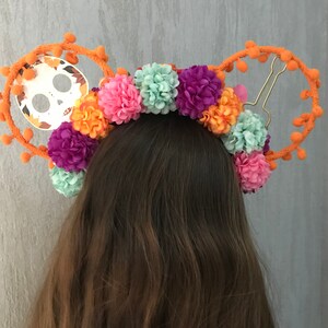 K&ouml;nnte beinhalten: Ein orangefarbenes Stirnband mit einem Totenkopfdesign und bunten Bommeln. Das Stirnband ist mit verschiedenen Blumen in den Farben Rosa, Lila, Orange und Gr&uuml;n verziert.